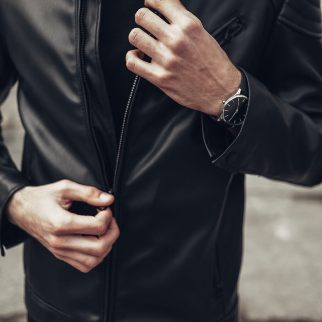 A close-up of a person wearing a black zip-up leather jacket, with hands adjusting the zipper. The jacket has a sleek, matte finish and a silver watch on the wrist.