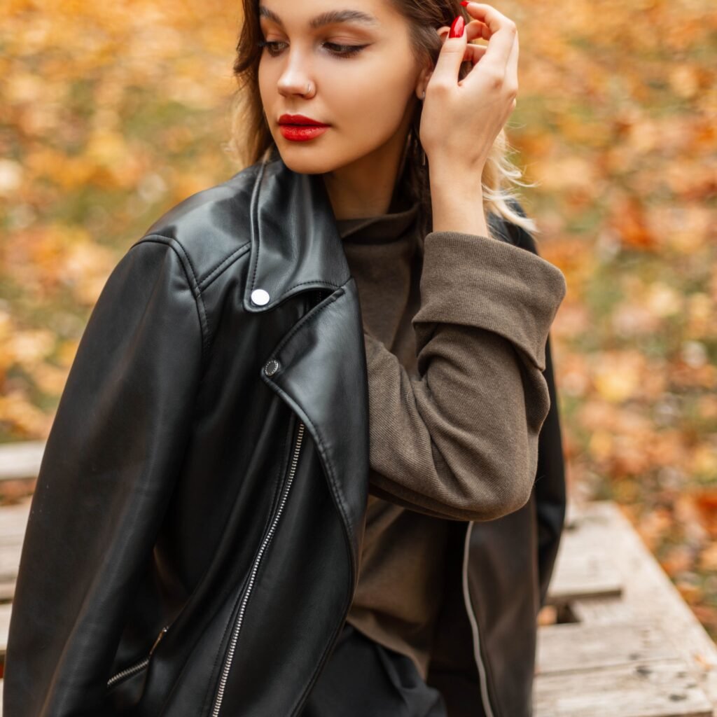 A stylish woman wearing a black zip-up leather jacket over a brown hoodie, standing outdoors with autumn leaves in the background.