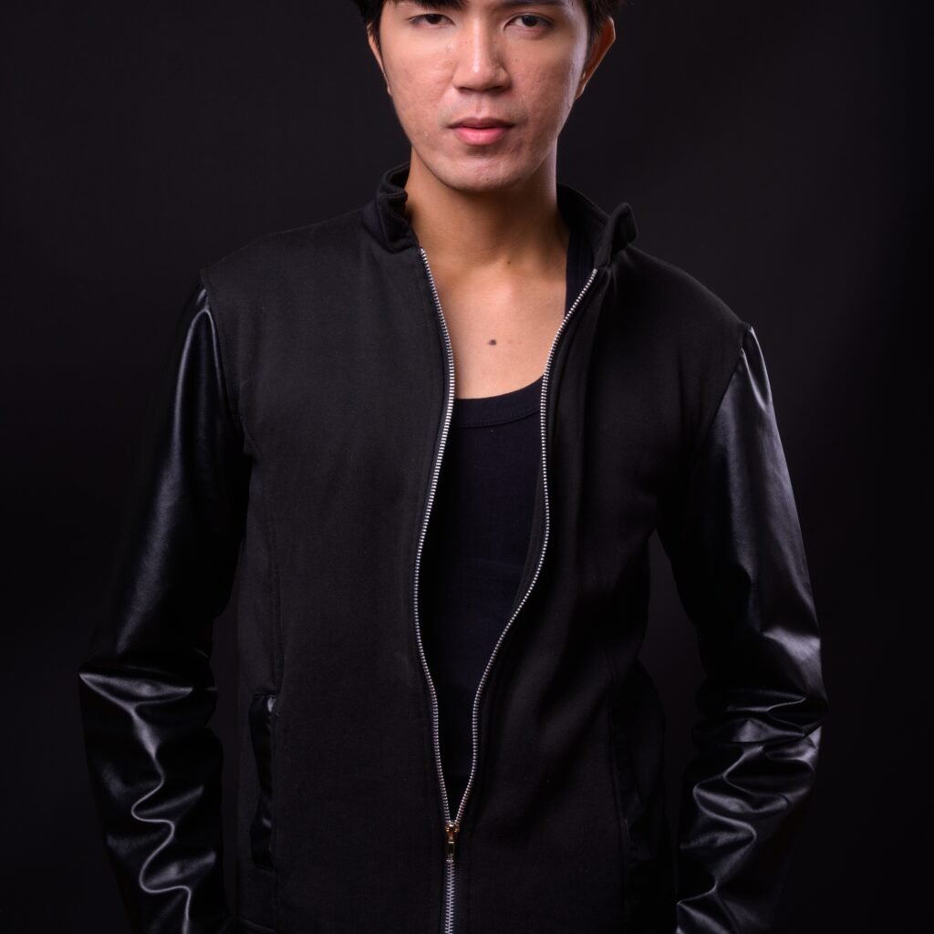 A person wearing a black zip-up leather-sleeve jacket against a dark background; the jacket is unzipped at the top, revealing a black shirt underneath.