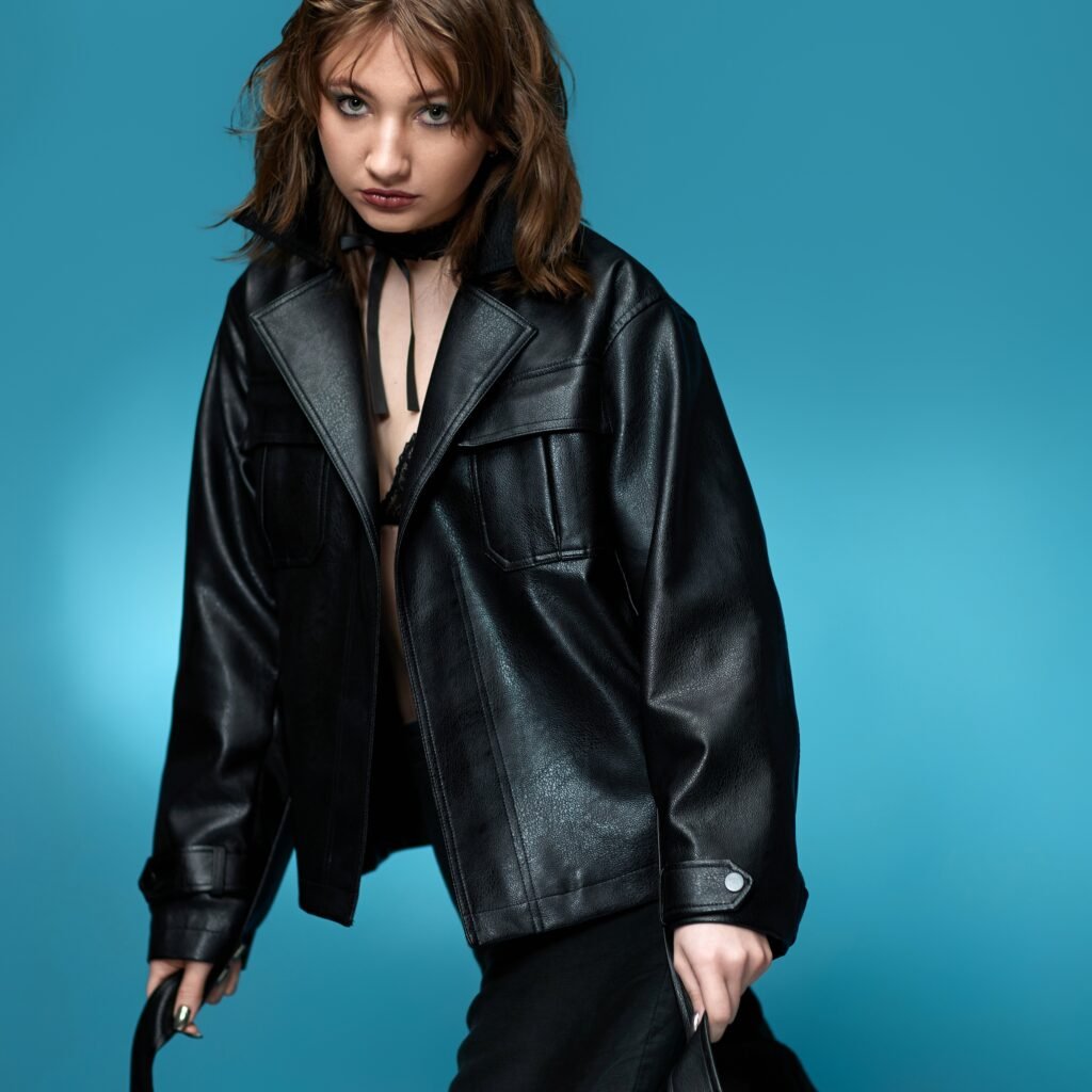 A young woman wearing a black leather zip-up jacket with wide lapels and a relaxed, edgy pose against a teal background.