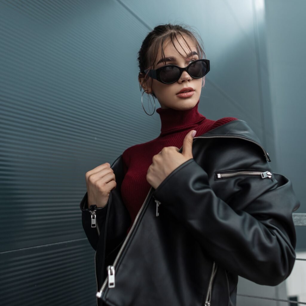 A stylish woman wearing a black zip-up leather jacket over a burgundy turtleneck, with oversized black sunglasses and hoop earrings. She stands against a blue-gray textured wall, hands gripping the jacket's lapels.Modern ZIP Up Leather Jacket