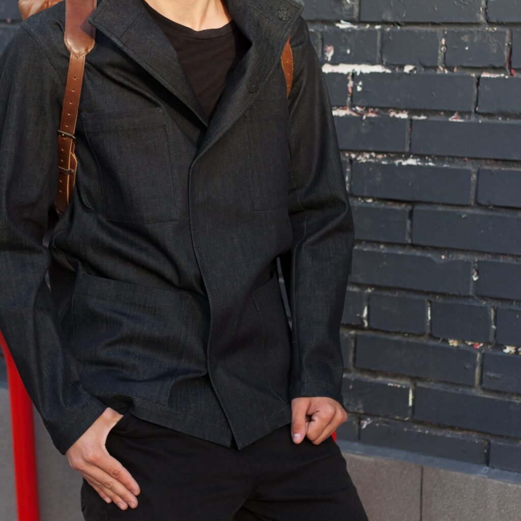 A person wearing a dark denim-like zip-up jacket with a high collar, black shirt, and a brown shoulder strap against a brick wall.Modern ZIP Up Leather Jacket