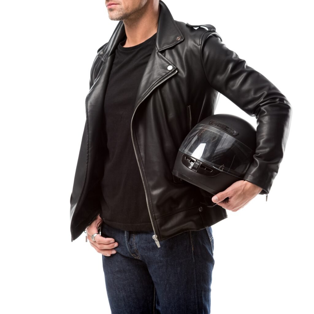A man wearing a black leather zip-up jacket holds a black full-face helmet under his arm. He’s dressed in a black shirt and dark jeans, posing against a white background.

