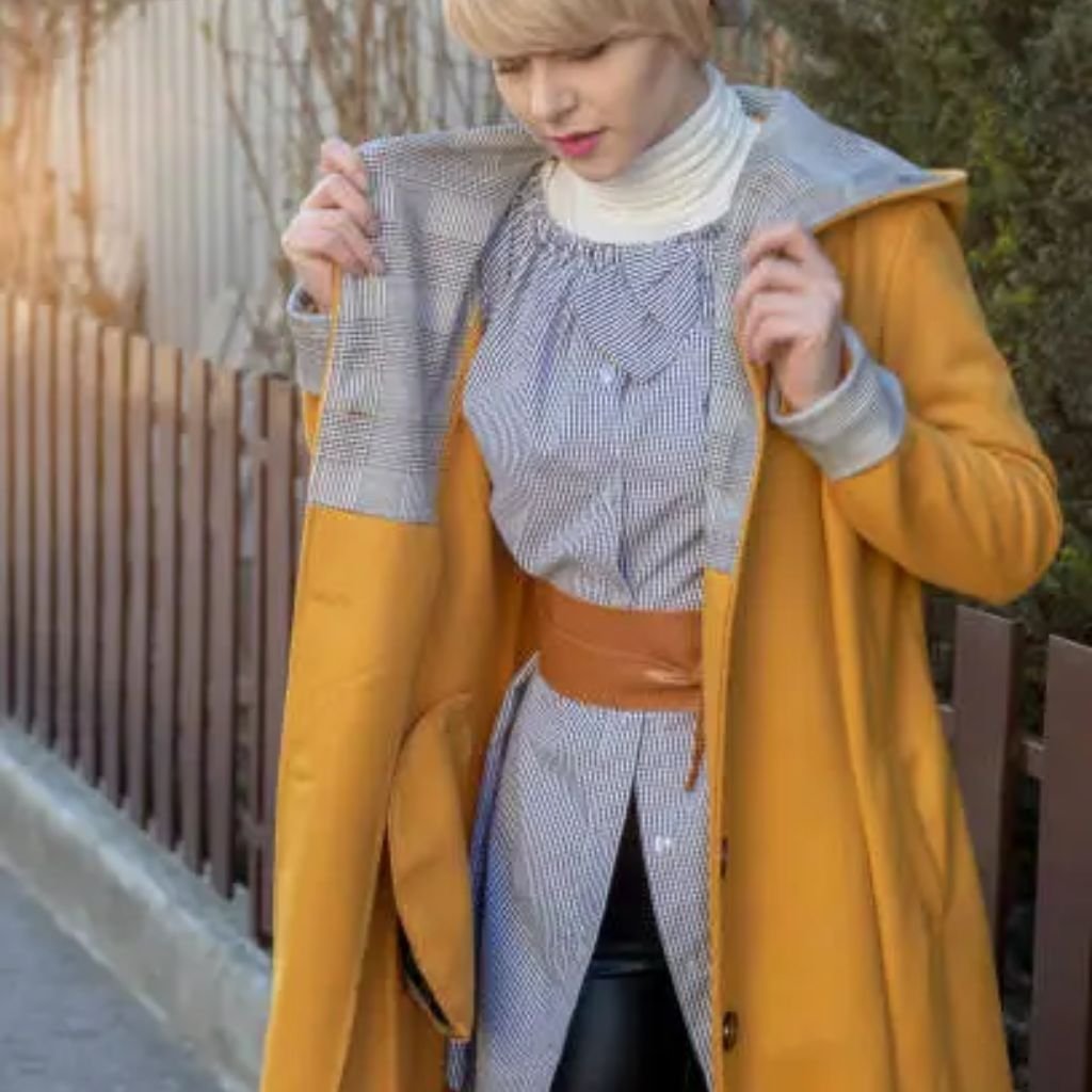 A person wearing a black baseball cap, black sleeveless top, and a black puffer jacket draped off one shoulder stands against a light-colored wall.Belted Winter Coats