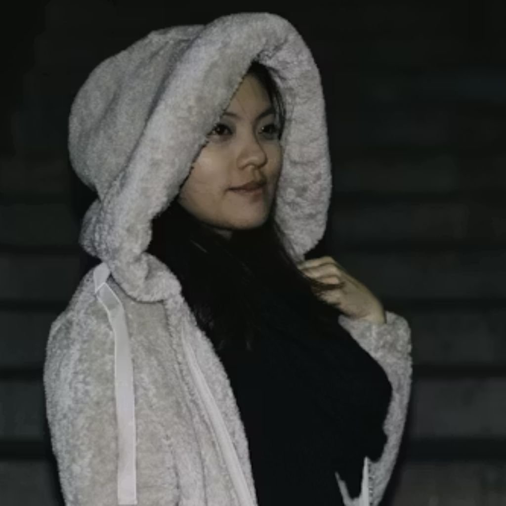 A person wearing a light-colored, fluffy hooded jacket stands in a dimly lit setting, looking to the side with a calm expression.