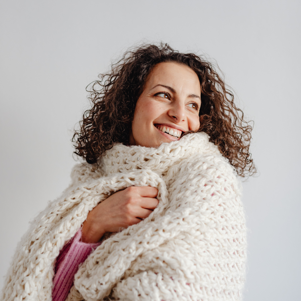A smiling woman with curly hair wrapped in a winter fabric which is thick, cream-colored knitted blanket, looking to her left.