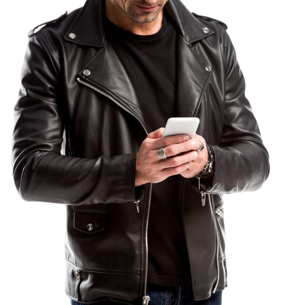 A stylish man wearing a black leather zip-up jacket, holding a white smartphone with both hands.
