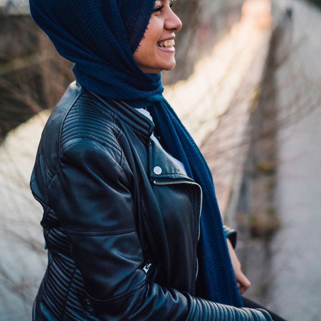 A person wearing a black leather jacket with quilted stitching and a detachable hood, accessorized with a long dark blue scarf. The individual is smiling and standing outdoors near a stone wall.
