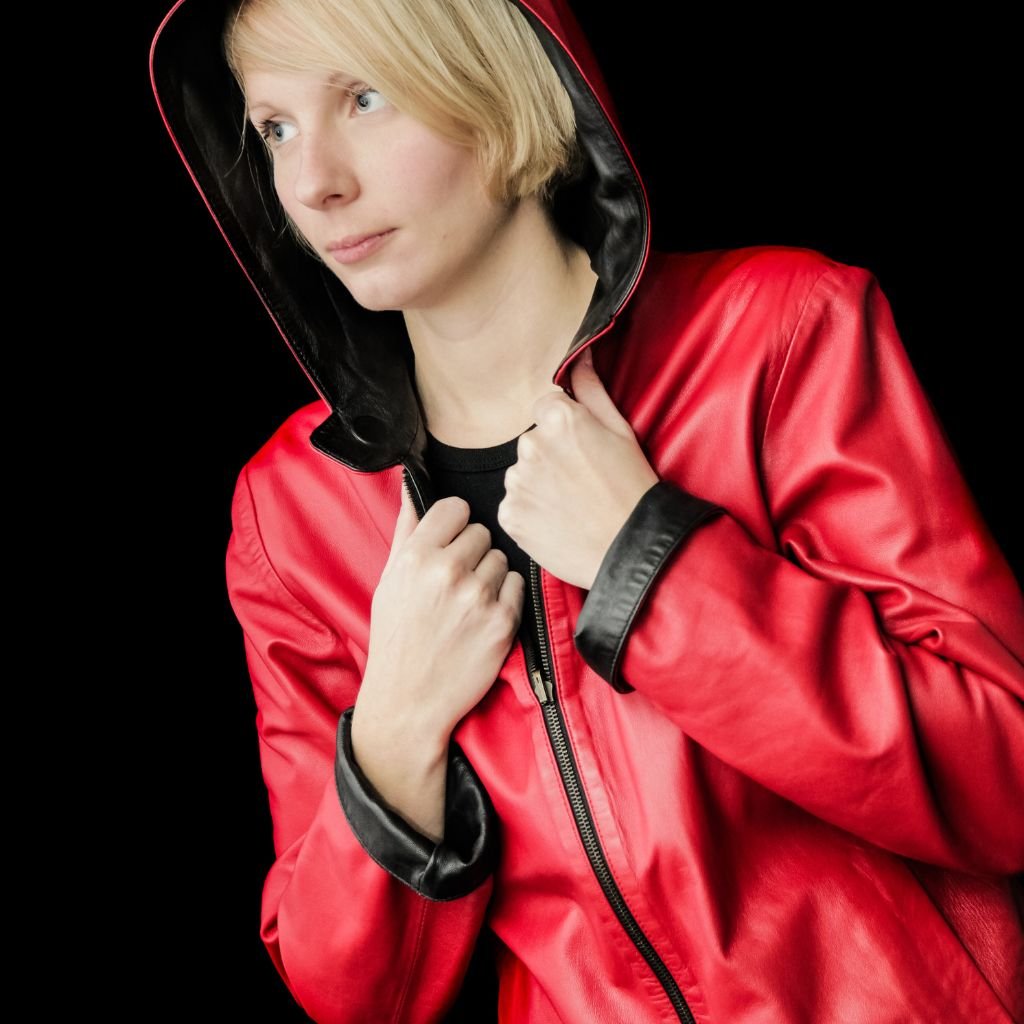 A person wearing a bright red hooded leather jacket with black lining, clutching the zipper with both hands. The background is black, and the person has light blonde hair and a calm expression.