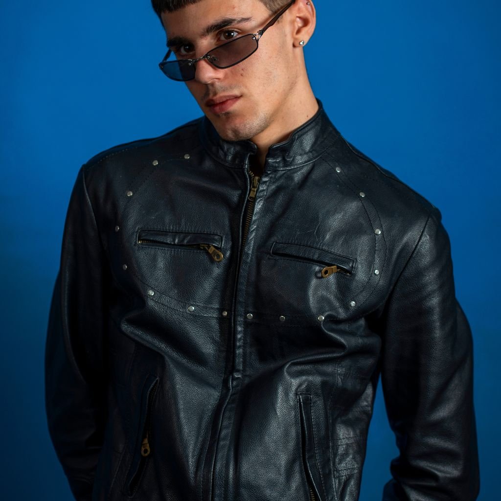 A stylish man wearing a black leather jacket with zip pockets and metal stud details stands against a blue background.