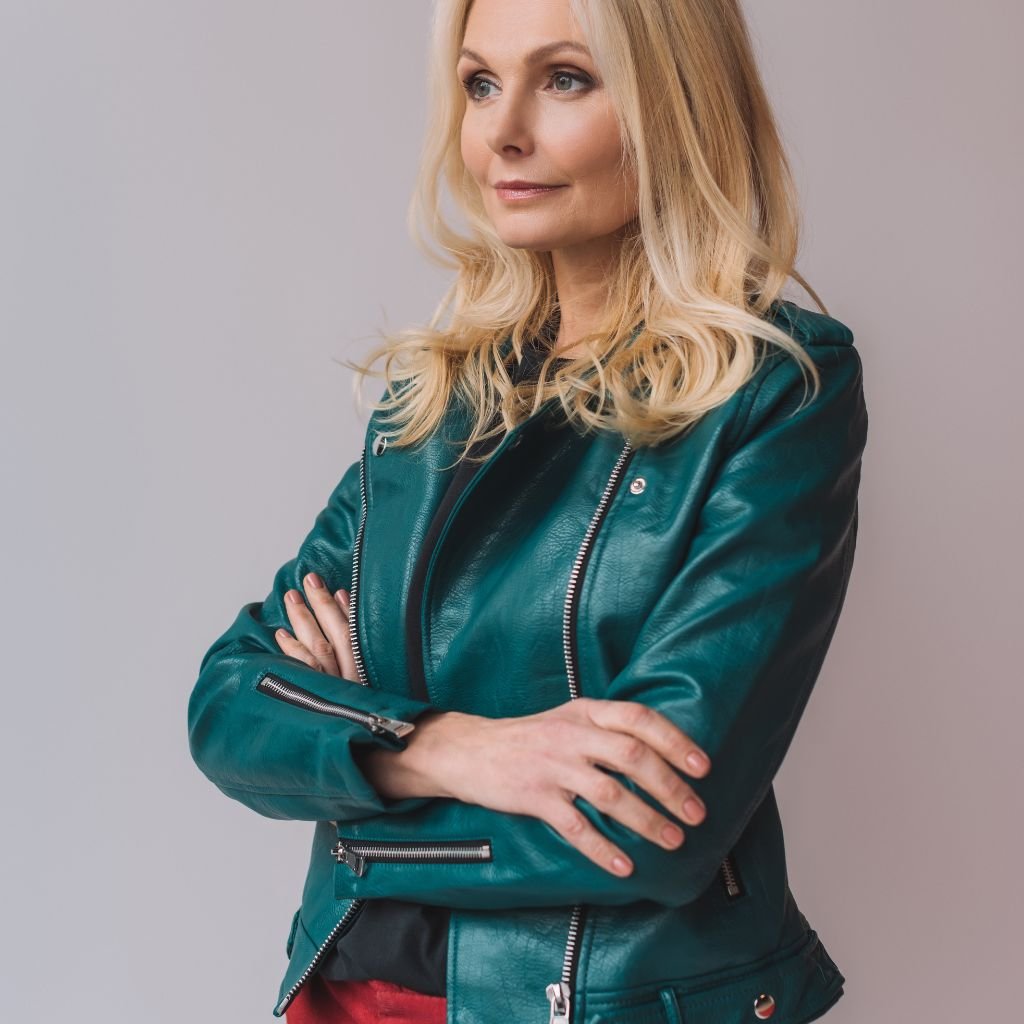 A stylish blonde woman wearing a teal leather fashion jacket with silver zippers, arms crossed, standing against a light gray background.