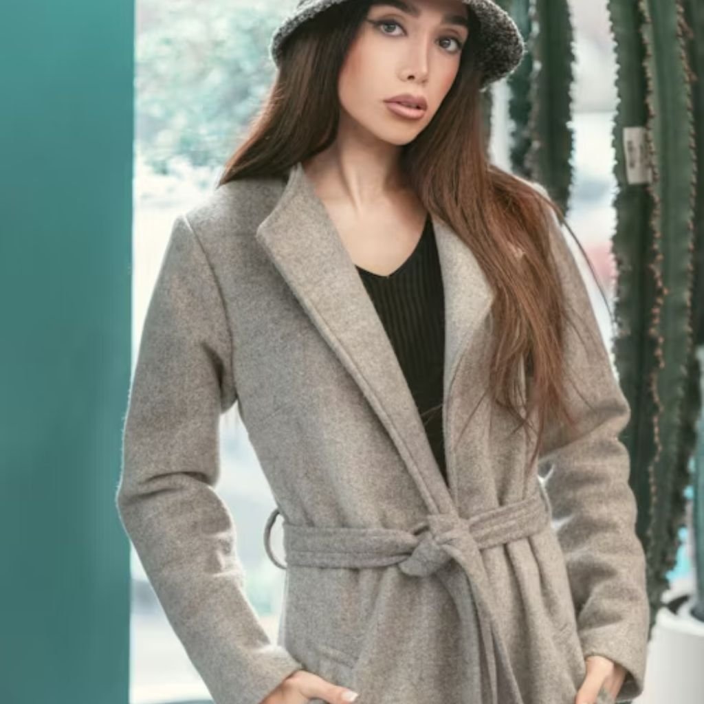 A person wearing a gray overcoat, black scarf, and sunglasses, standing against a dark background and adjusting their coat.The Wool Peacoat Comeback: