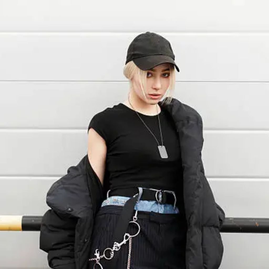 A person wearing a black baseball cap, black sleeveless top, and a black puffer jacket draped off one shoulder stands against a light-colored wall.Belted Winter Coats