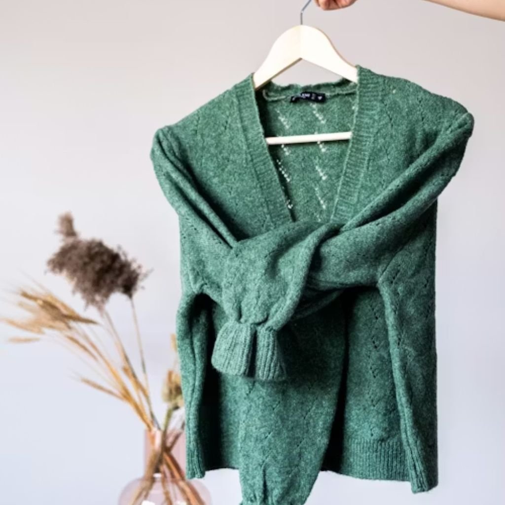 Green knitted V-neck sweater hanging on a wooden hanger against a light background with dried plants.Is Pure Wool Worth It?