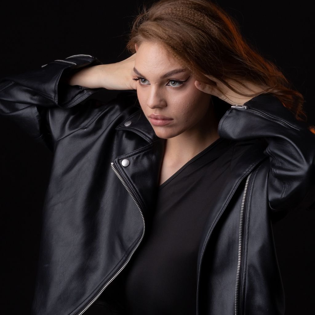 A stylish woman wearing a black leather jacket, posed with hands near her head against a dark background.
