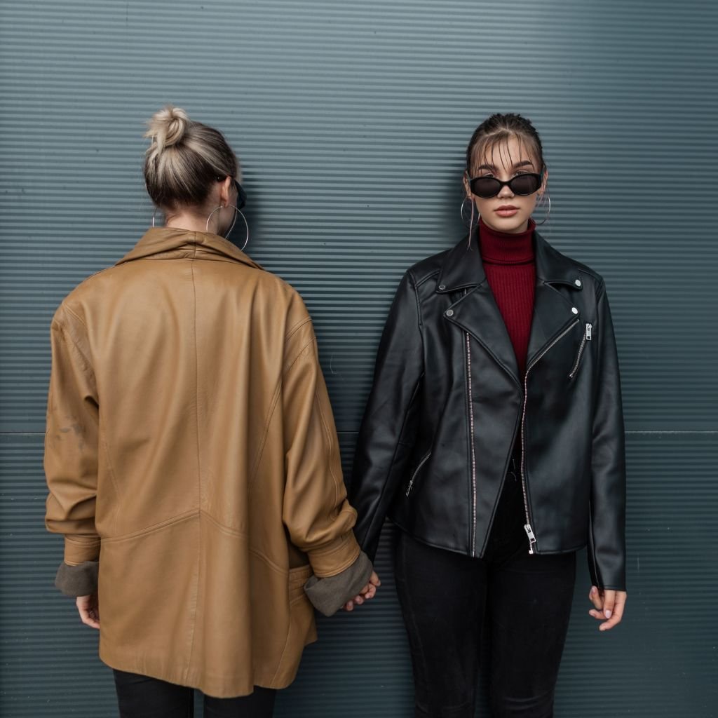 Two stylish women stand against a ribbed metal wall; one faces away wearing a tan leather jacket with rolled cuffs, the other faces forward in a black leather jacket over a red turtleneck, wearing sunglasses and hoop earrings.

