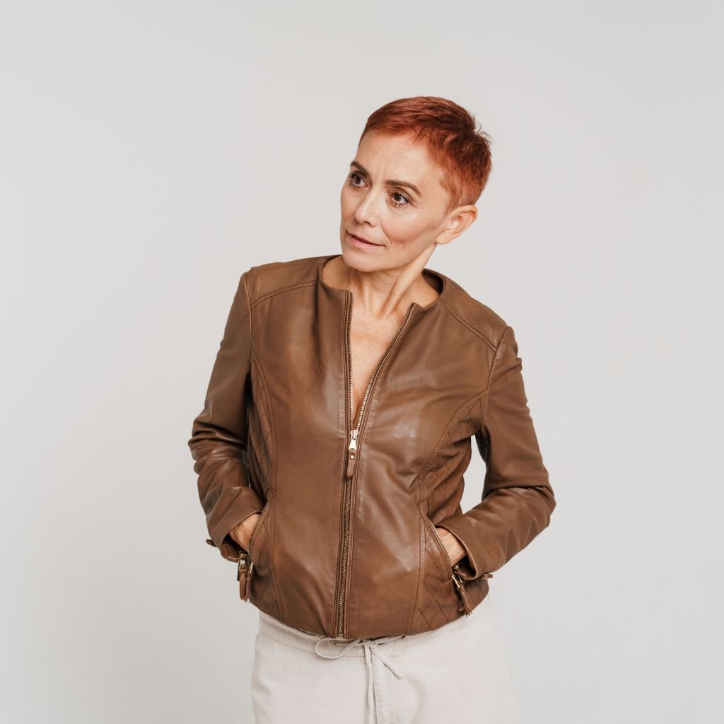 A woman with short red hair wearing a brown leather jacket, standing with hands in pockets against a light gray background.
