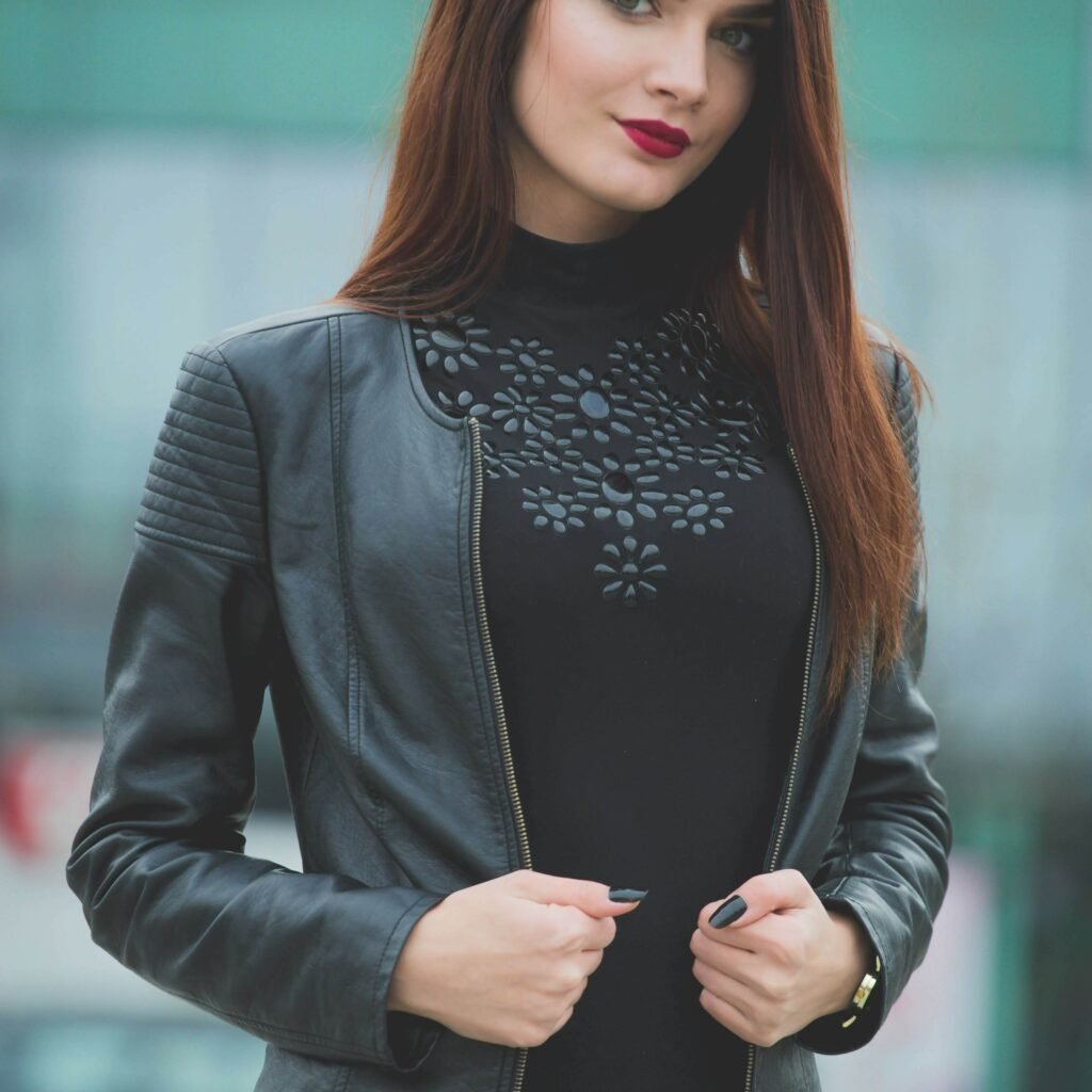 A stylish woman wearing a black leather blazer with quilted shoulders over a black top, standing outdoors with a soft green background.
