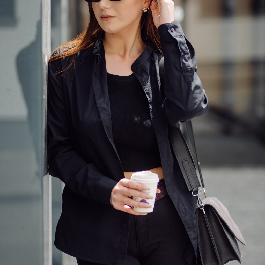 A stylish woman stands outdoors wearing a black leather blazer over a black top, holding a white disposable coffee cup with both hands. She has dark sunglasses and a black crossbody bag.

