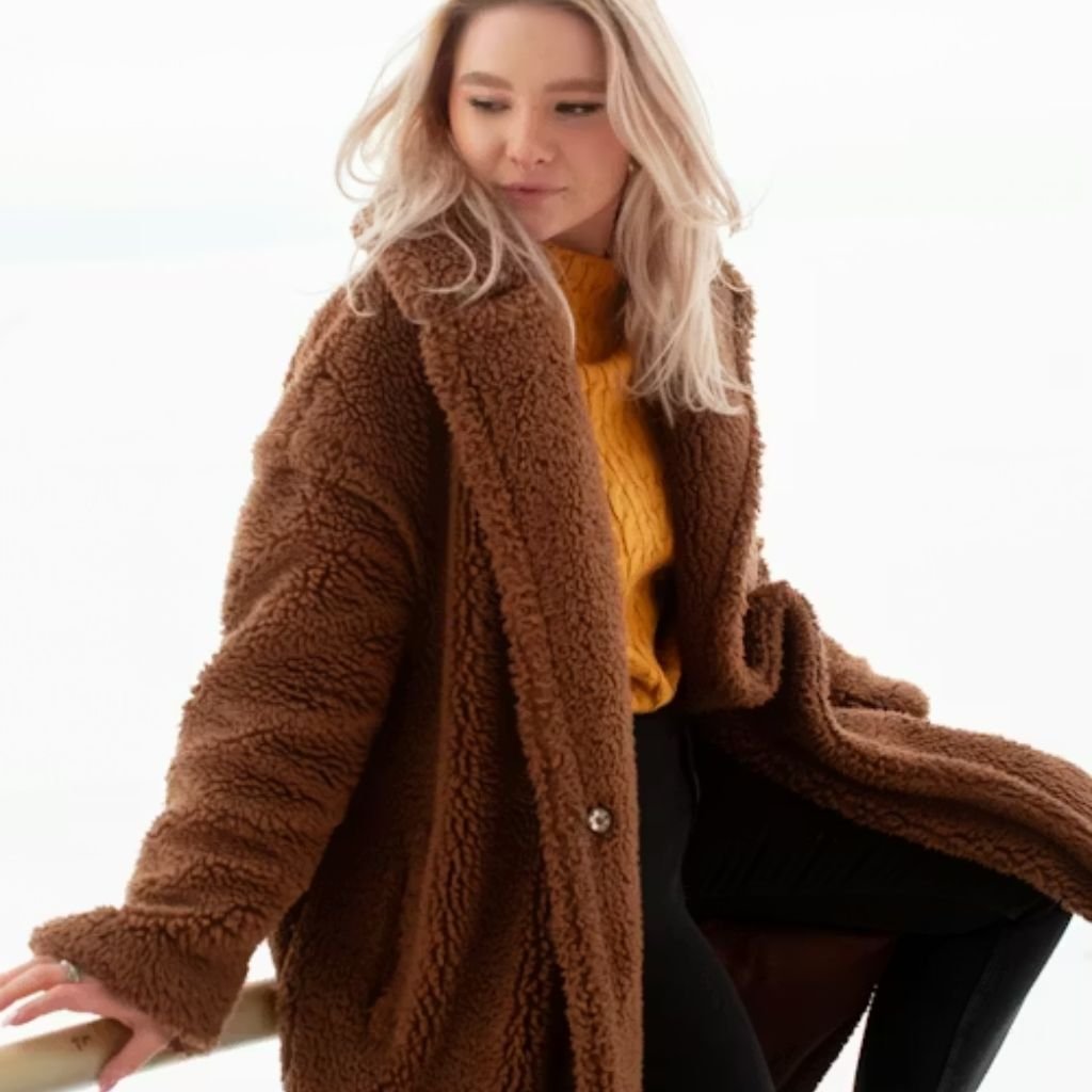 A person wearing round sunglasses and a light-colored coat stands on a bridge with a city skyline in the background.The Wool Peacoat Comeback: