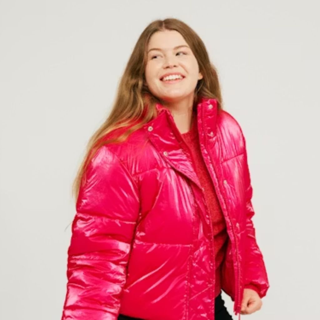 A smiling woman wearing a black quilted hooded jacket stands with her hands adjusting the collar, posing confidently against a plain white background.Why Oversized Padded Jackets