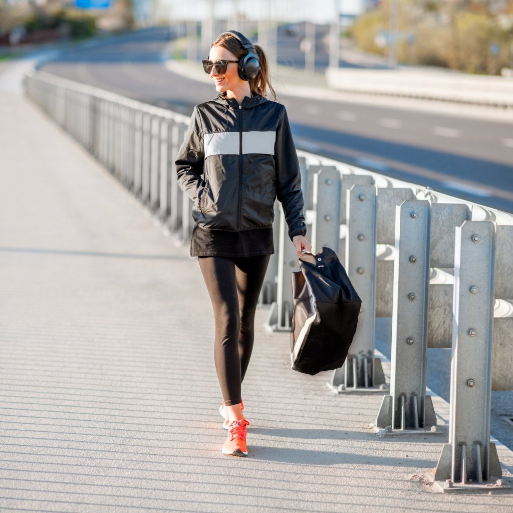 A woman walking on a sidewalk beside a metal railing, wearing a black windbreaker with a light gray stripe, black leggings, bright pink sneakers, sunglasses, and headphones, carrying a black tote bag.