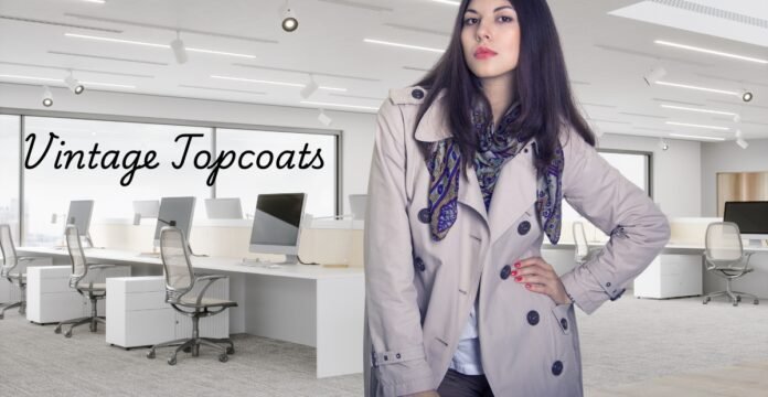 A woman wearing a light trench coat and scarf standing in a modern open-plan office with white desks and chairs; ‘Vintage Topcoats’ text on the window. Vintage Topcoats