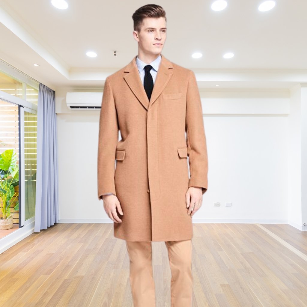A man stands indoors wearing a camel-colored overcoat over a white shirt and black tie; he faces forward with a neutral expression. The room has wooden flooring, white walls, and bright ceiling lights.