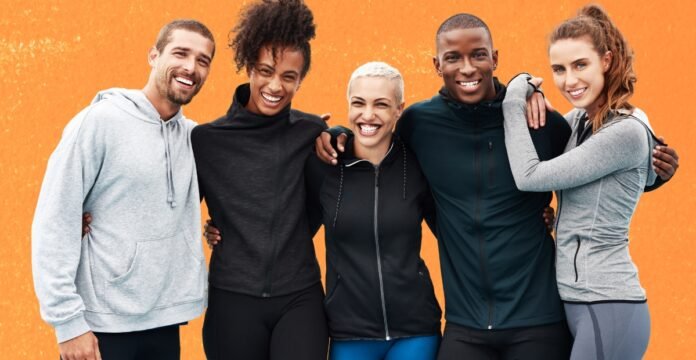A diverse group of five friends stands arm-in-arm against an orange wall, smiling at the camera. They’re dressed in athletic wear. What Is Track Jacket