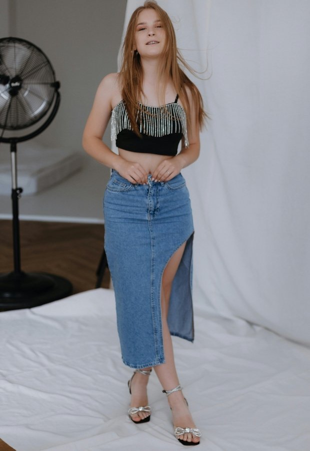 A young woman posing in a studio wearing a black beaded crop top, a light-wash denim A-line skirt with a high waist and a front slit, and silver strappy heels. A large studio fan is visible in the background.