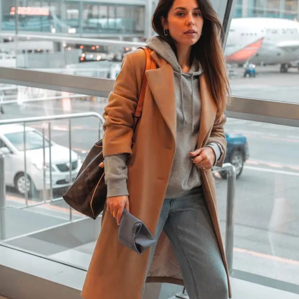A stylish woman walking in an airport terminal, wearing a camel trench coat over a gray hoodie, light-wash jeans, and carrying a gray clutch and a large brown tote bag.