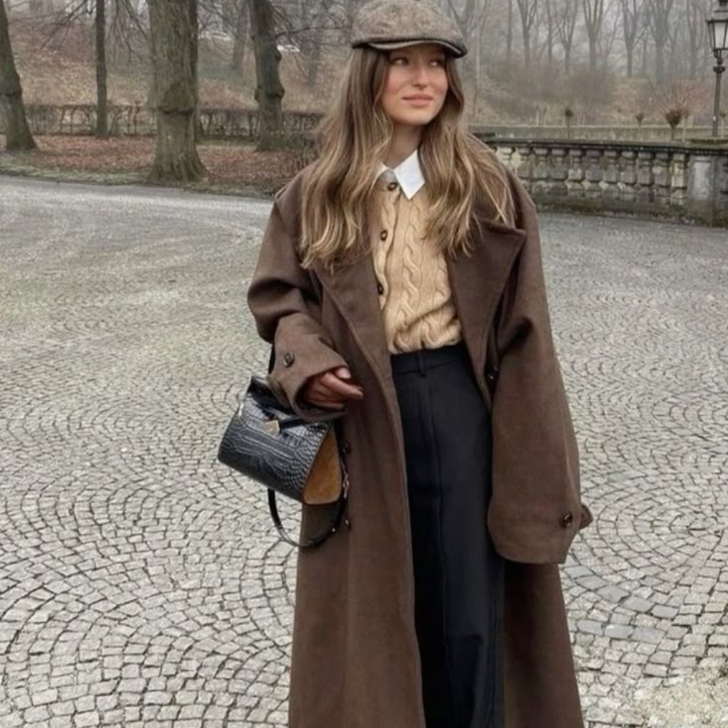 A stylish woman stands on a cobblestone path wearing a long brown coat, a beige cable-knit sweater, black wide-leg trousers, a white collared shirt, a brown flat cap, and a small patterned handbag.