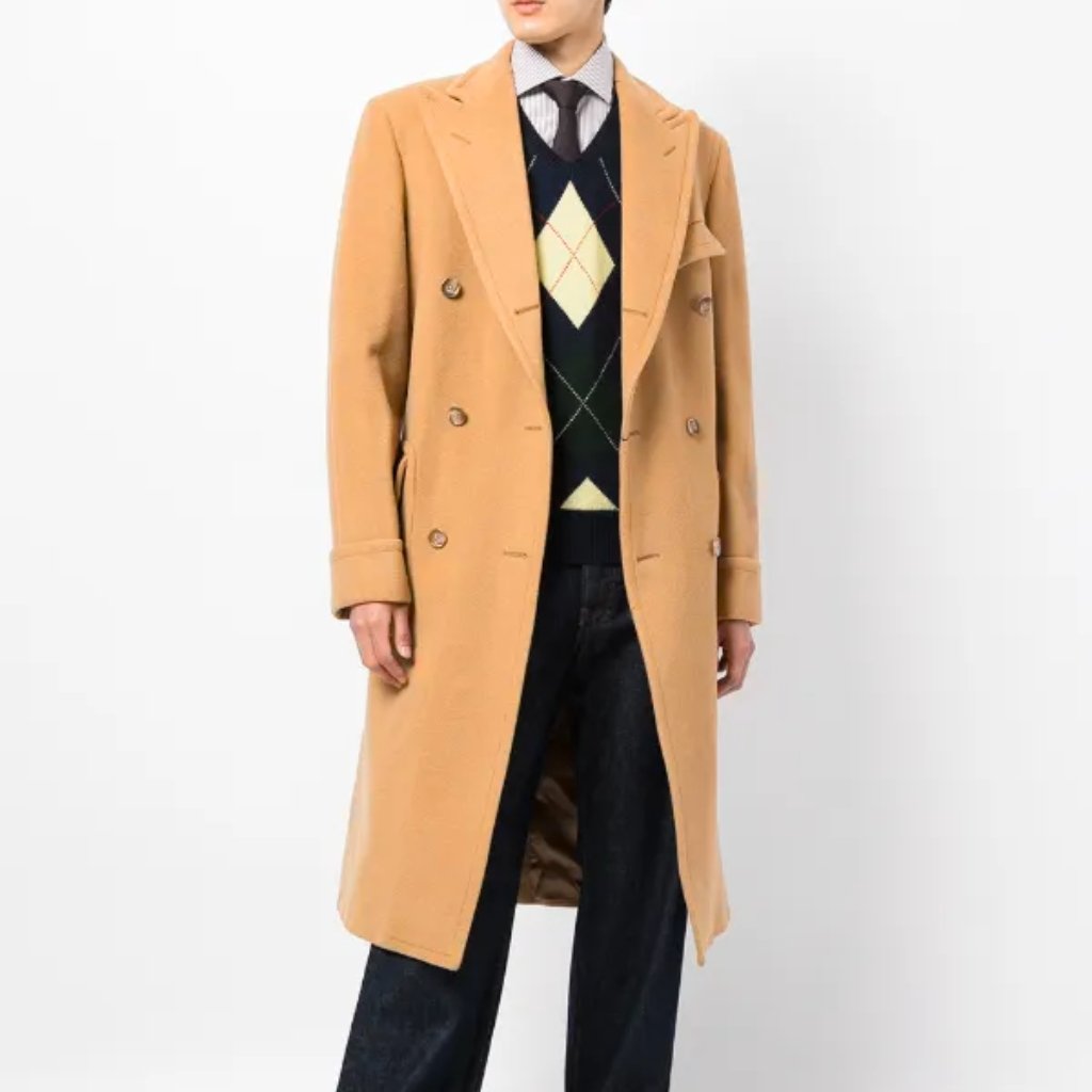 A person wearing a camel-colored double-breasted overcoat over a navy argyle sweater vest and white shirt with a tie, paired with dark jeans.