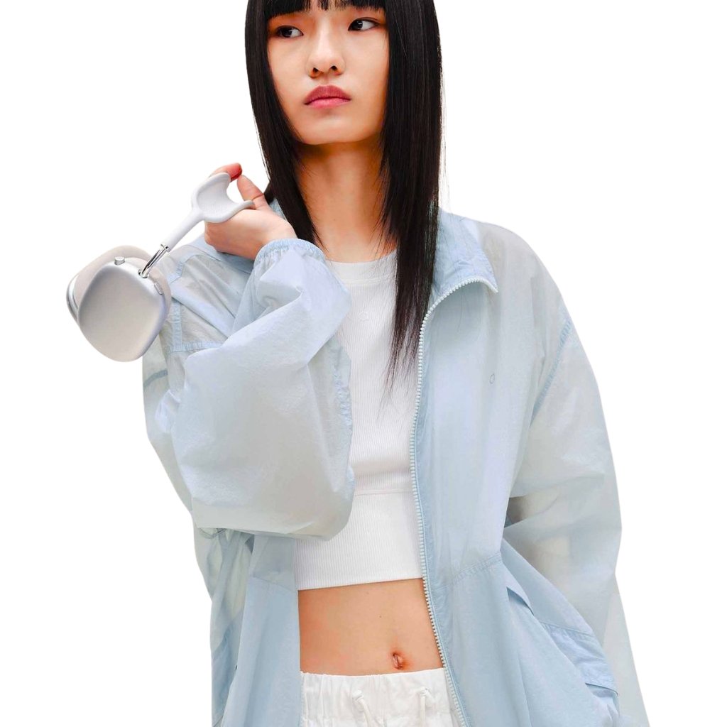 A young woman wearing a light blue windbreaker and white crop top, holding a handheld milk frother near her shoulder, looking off to the side with a neutral expression.