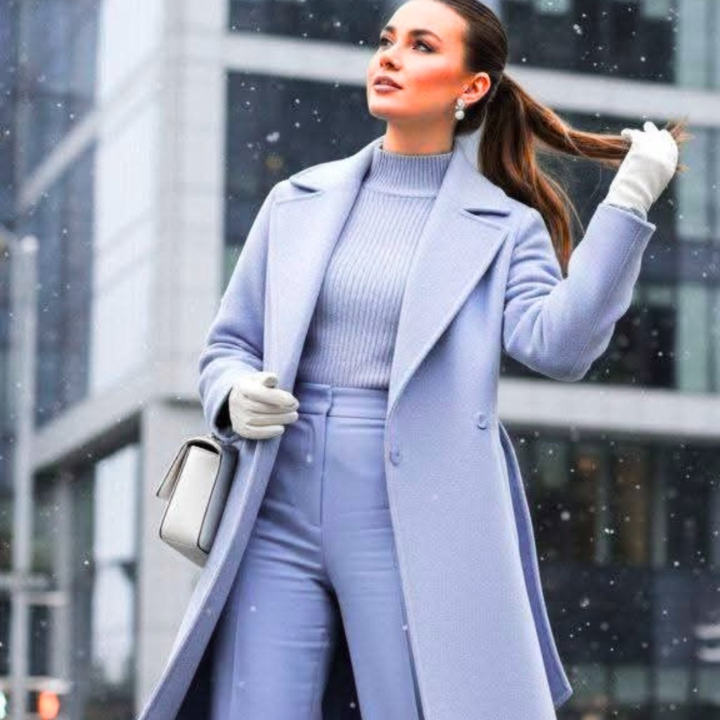 Woman walking in a snowy urban setting wearing a light lavender coat, matching sweater and pants, white gloves, and a white handbag.