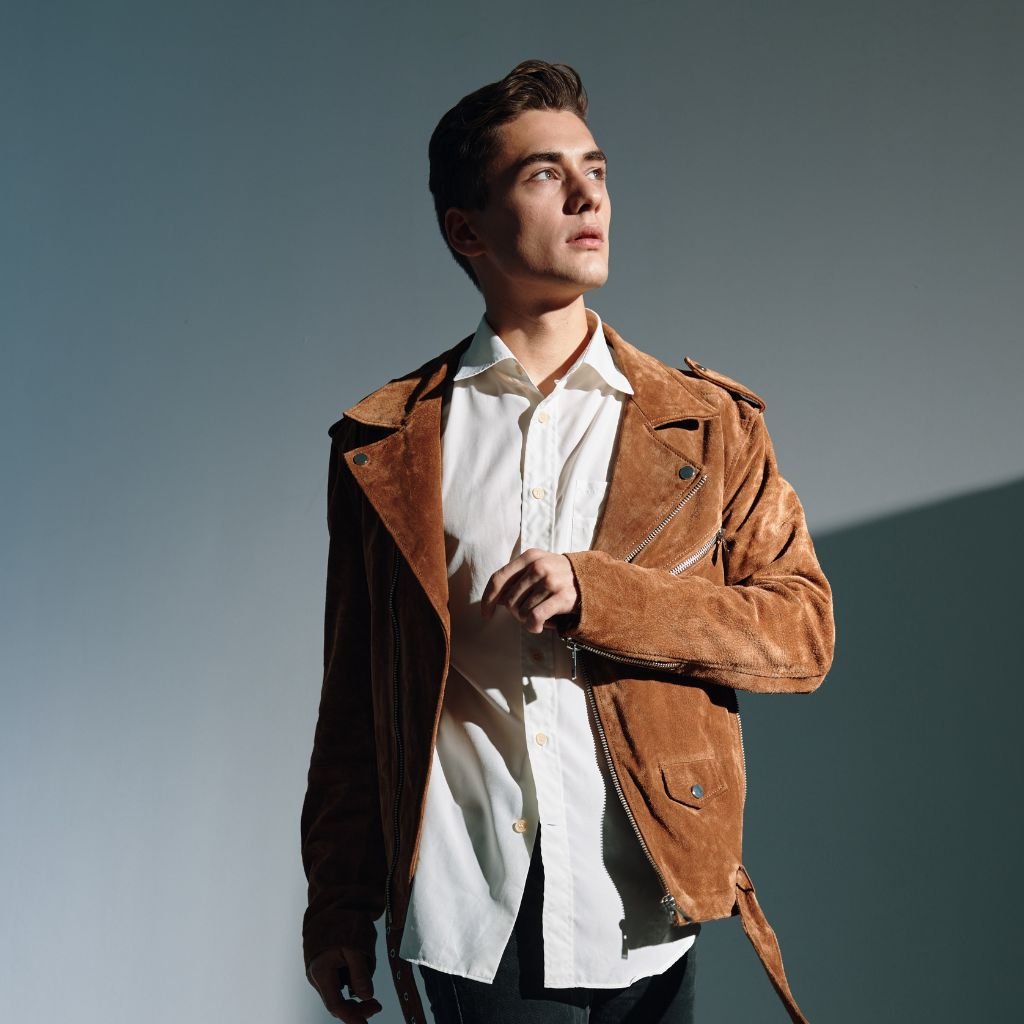 A young man stands against a neutral background, wearing a light beige shirt under a brown suede jacket with zippers and pockets, looking off to the side with a thoughtful expression.

