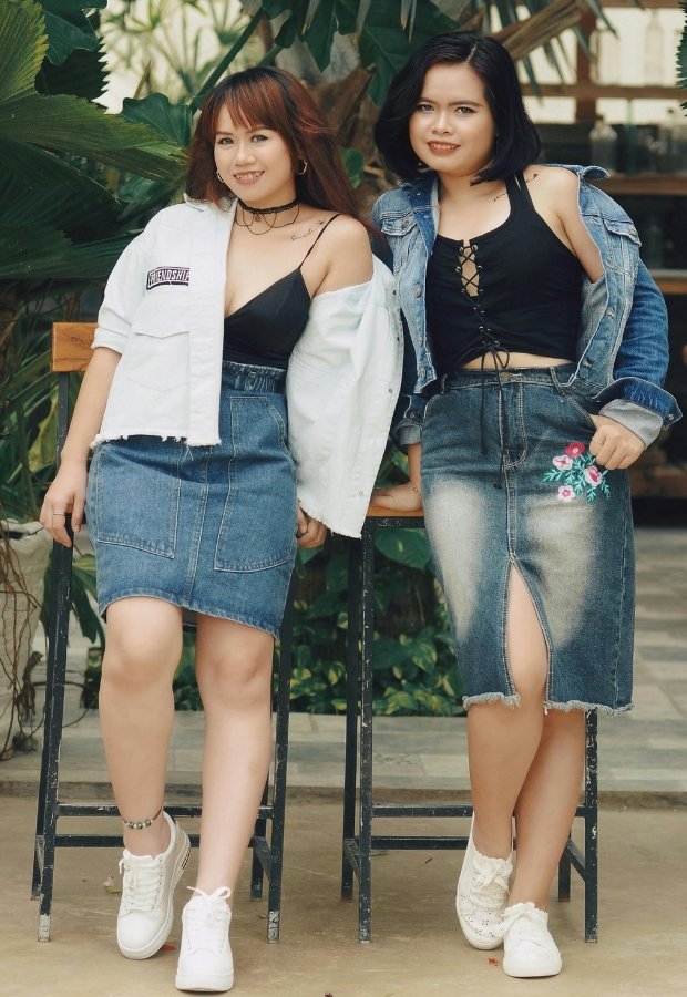 Two women standing together outdoors, both wearing denim skirts and black tops. The woman on the left wears a light denim A-line mini skirt with a white cropped jacket, white sneakers, and a choker; the woman on the right wears a mid-length denim A-line skirt with a front slit, a black lace-up top, a denim jacket draped over her shoulders, and white sneakers. They are smiling and leaning against a railing with greenery in the background.