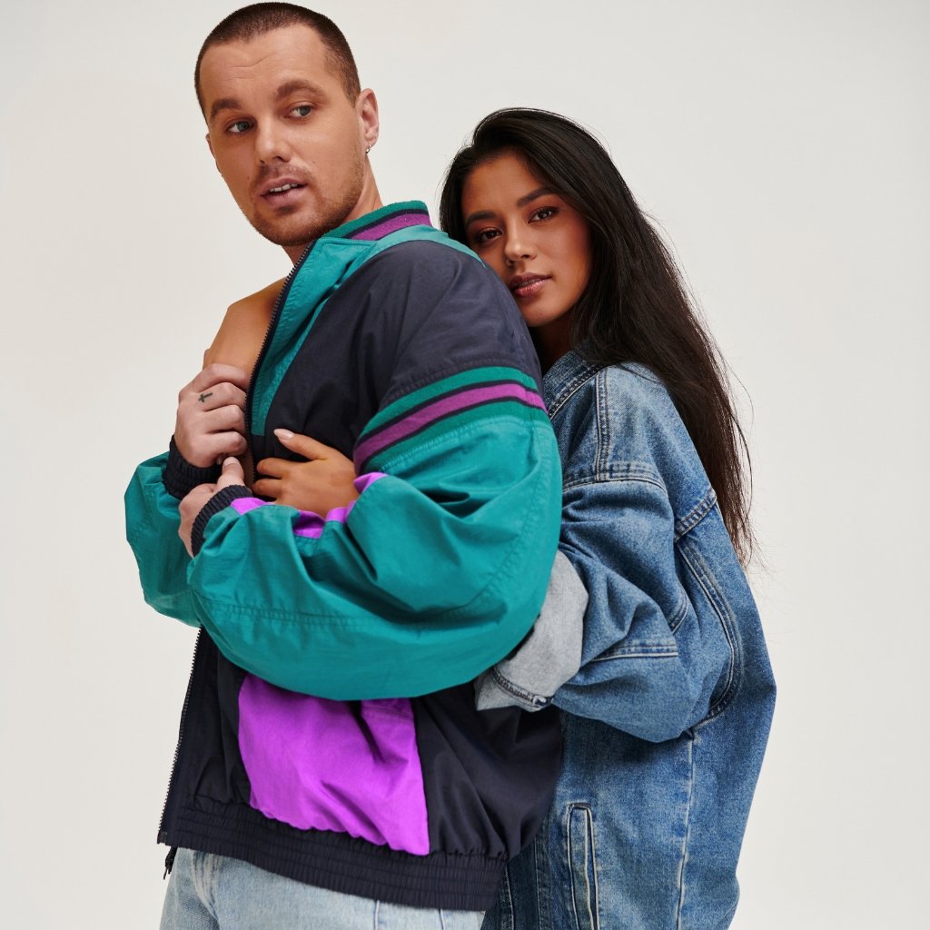 A young man wearing a colorful windbreaker stands in front, while a young woman in a blue denim jacket embraces him from behind, both looking off to the side against a light background.