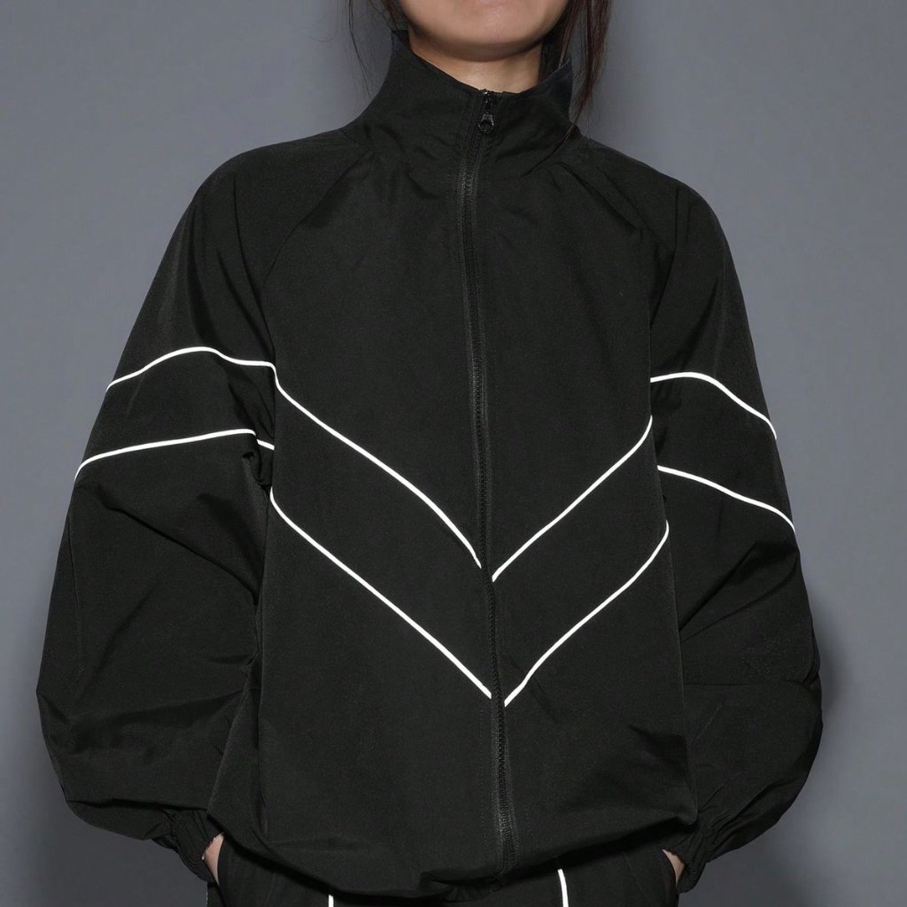A person wearing a black zip-up jacket with white chevron-like lines across the chest and arms. The jacket has a high collar and puffed sleeves, with hands in pockets.