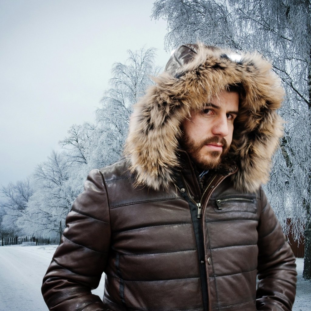 A man wearing a brown leather winter coat with a large fur-lined hood stands outside on a snowy, tree-lined street.Classic Leather Parka Jacket