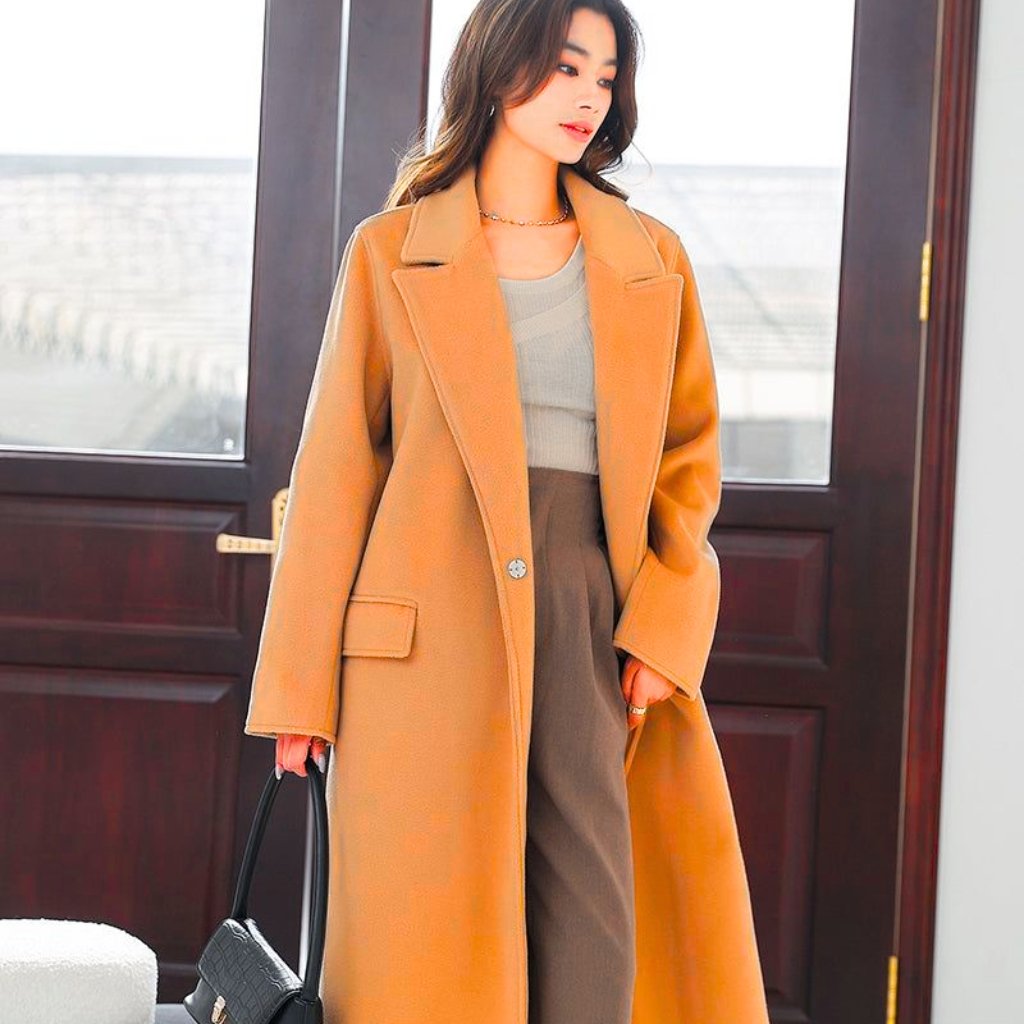 A stylish woman stands indoors wearing a camel-colored long wool coat over a light beige top and taupe tailored pants. She carries a small black handbag and wears a delicate necklace, with her hair down and natural makeup. The background shows a dark wooden door and large windows letting in daylight.