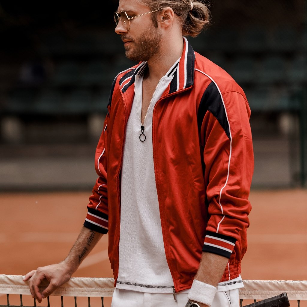 A stylish man leaning on a tennis net, wearing a red track jacket over a white polo, with sunglasses and a man bun.