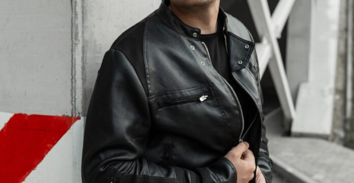 A person wearing a black leather field jacket with a zipper, standing against a concrete wall with red diagonal markings.