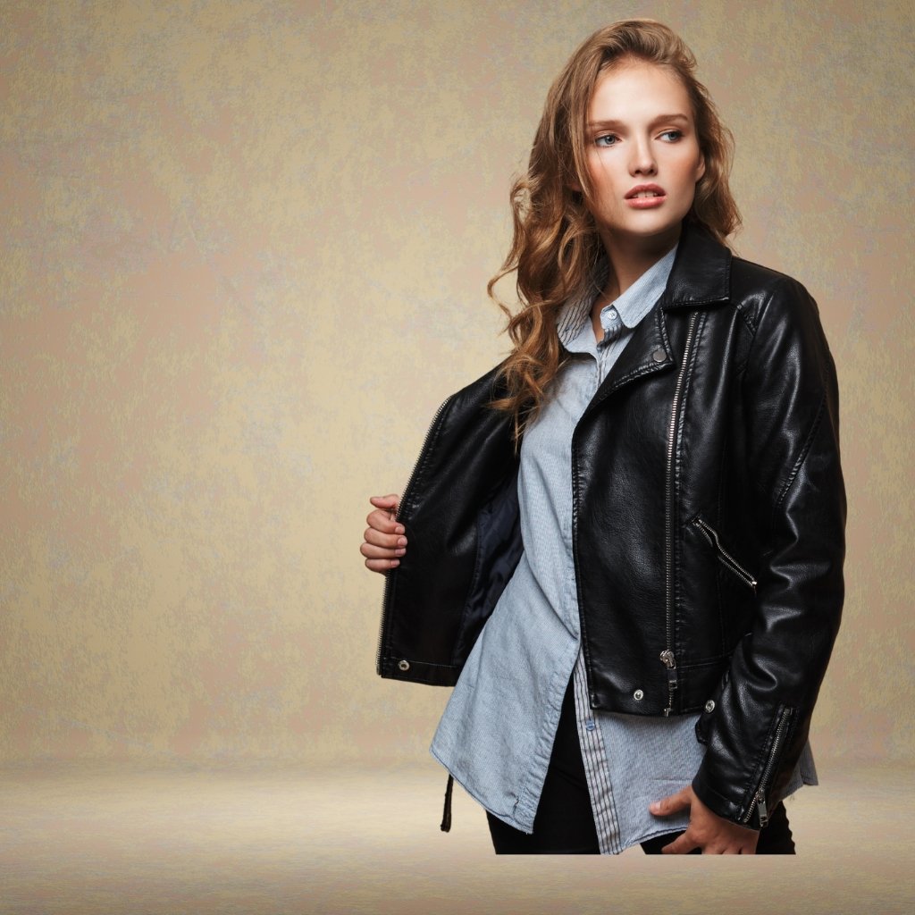 A stylish young woman wearing a black leather short jacket over a light blue button-down shirt, posing confidently with one hand on the jacket. The background is a soft textured beige.