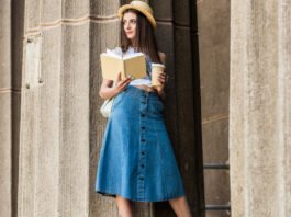 A stylish young woman wearing a straw hat and a light-colored top, leaning against a textured stone wall while holding a book and a takeaway coffee cup; she is wearing a denim A-line skirt.