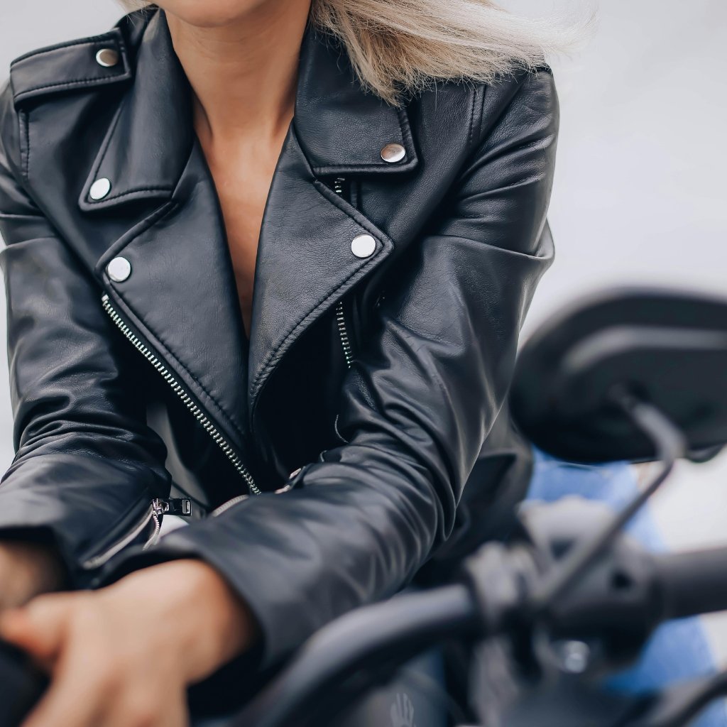 A close-up of a person wearing a black leather motorcycle jacket with silver snaps and zippers, sitting on a motorcycle.