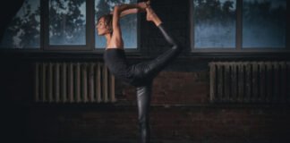 Yoga Leather Pant: Comfort, Style, and Flexibility Combined A person performing a yoga pose indoors. They stand on one leg while bending the other leg upward, reaching back to hold the foot with both hands. The setting includes brick walls and large windows with a dark, moody atmosphere. The person wears a black leather-like yoga pant and a sleeveless top.
