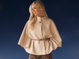 A stylish woman wearing a beige cape jacket with wide, flowing sleeves and a high collar. The cape is belted at the waist with a thin belt, and she is standing against a dark blue background.