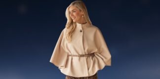 Cape Jacket for Women: Timeless Elegance Meet Modern Style A stylish woman wearing a beige cape jacket with wide, flowing sleeves and a high collar. The cape is belted at the waist with a thin belt, and she is standing against a dark blue background.