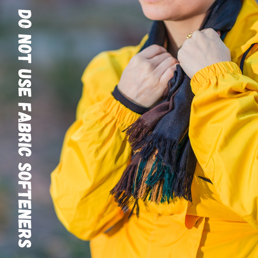 A person wearing a bright yellow rain jacket holds a dark fringed scarf close to their neck, outdoors.